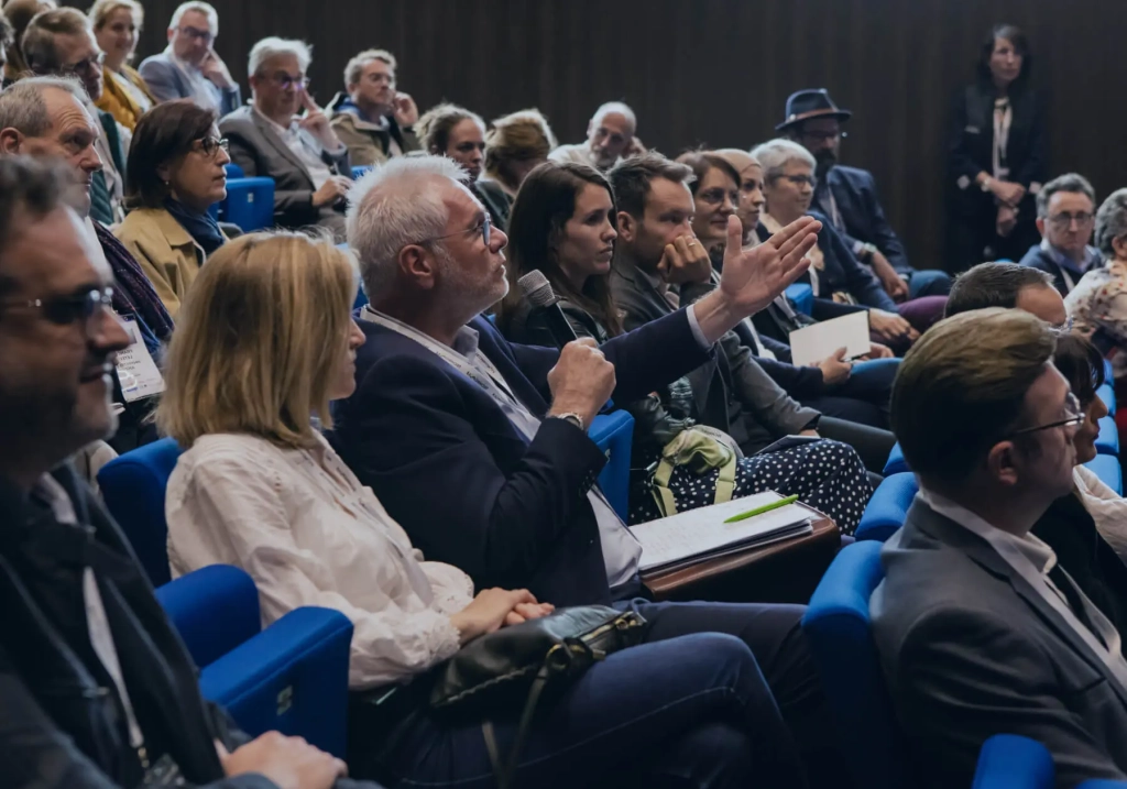 Un homme assis dans un amphithéâtre au milieu de plusieurs personnes qui parle dans un micro